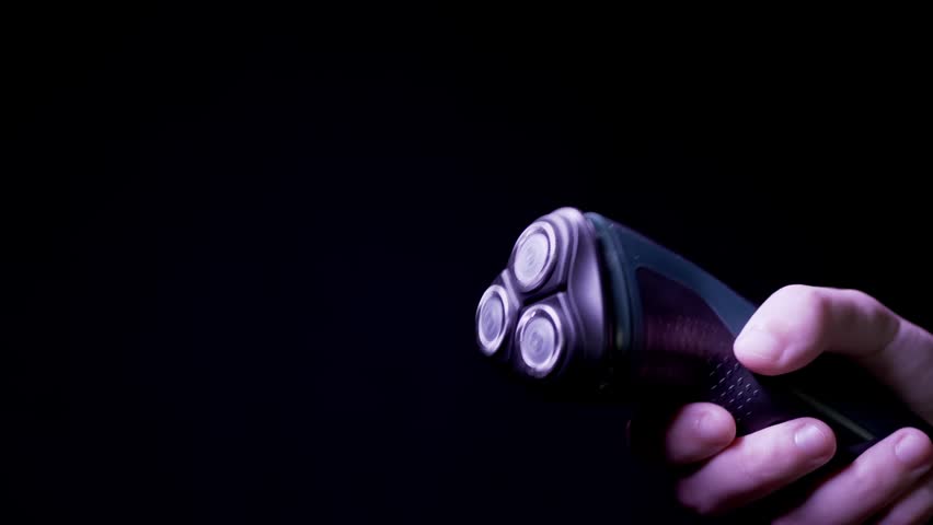 Close-up view of a hand holding a black electric rotary shaver and turning it on against a dark background. Side view. Concept of daily hygiene routine, modern grooming technology, and