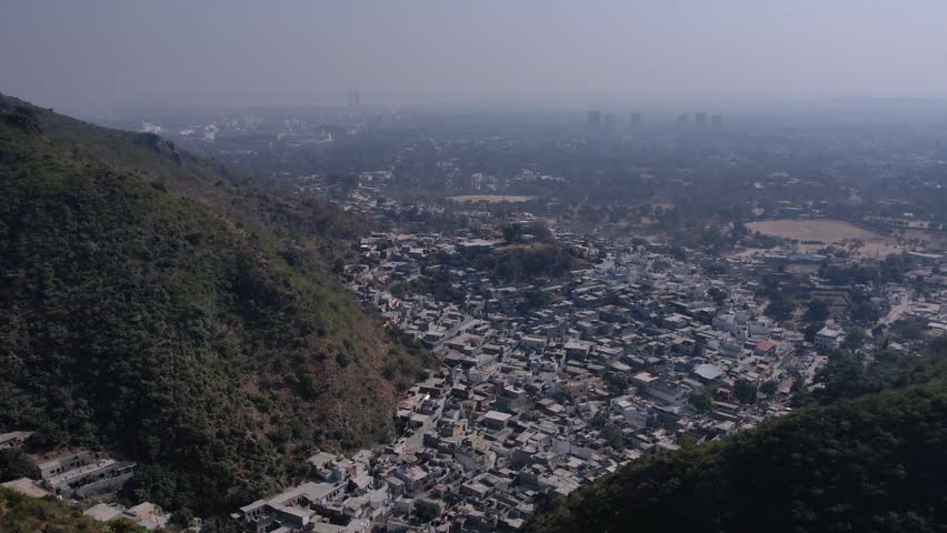 Filmed on February 9, 2025, at Saidpur Village, Margalla Hills, Islamabad, Pakistan. Drone aerial shot panning over a densely packed hillside settlement surrounded by forested mountain slopes, with an expansive city sprawl and distant skyscrapers fading into haze. Aerial city view, hillside houses, mountain settlement, urban landscape, city from above, rooftop view, mountain city, wide aerial panorama.