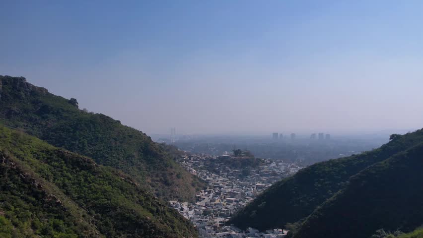 Saidpur Village - 9 February 2025, at Margalla Hills, Islamabad, Pakistan. Aerial drone footage of a dense hillside village nestled between green mountain ridges with a vast city skyline and high-rise buildings visible in the hazy distance. Mountain village aerial view, urban settlement, city skyline, green hills, residential area, cityscape from mountains, hazy atmosphere, drone panoramic.
