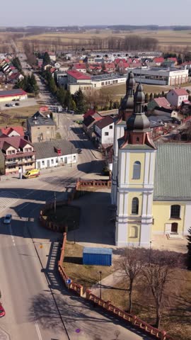 Vertical screen Panoramic Aerial View Stawiski Church And Town Center, Cars Parked Near Square, Surrounding Rooftops And Farmland Stretching To Horizon, Clear Midday Light And Calm Streets