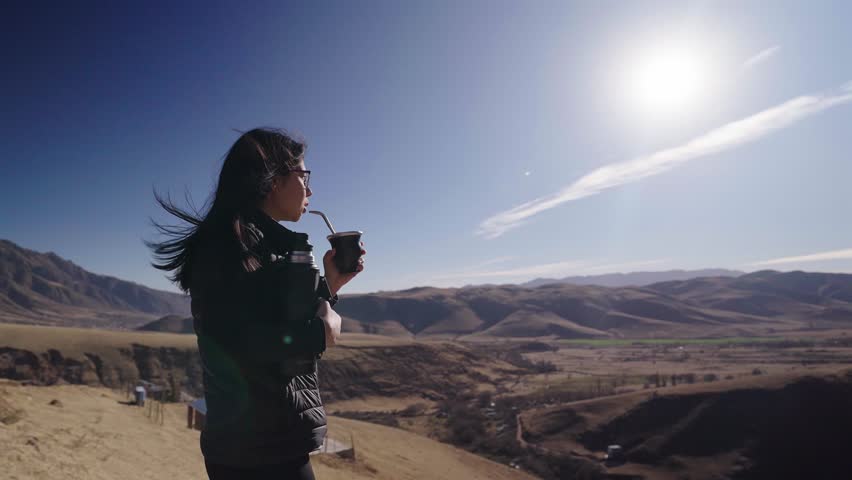 Woman drinking traditional mate tea while observing mountain valley landscape in northern Argentina showing relaxation, travel lifestyle and cultural connection with nature, slow motion side back shot