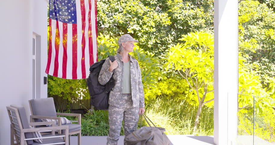 Woman pausing scanning walking toward camera on porch with US flag carrying duffel rucksack in camo. Female, military, camouflage, backpack, duffel, patio, outdoor