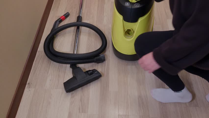 Person holding hose while assembling yellow, black vacuum cleaner on light wood floor, domestic cleaning preparation.