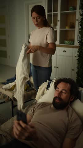 Stressed woman tidying up clothes while her lazy husband ignores her, using his phone. She gets angry, shouts, and starts a fight