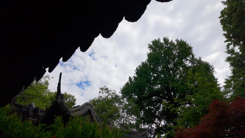Clouds drift above traditional rooftop and lush trees in an outdoor garden setting