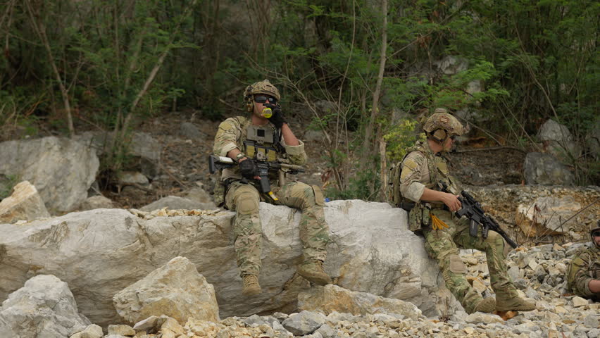 Two soldiers in full combat gear take a break to rehydrate while on a military operation in a rugged, rocky environment, showcasing tactical downtime.