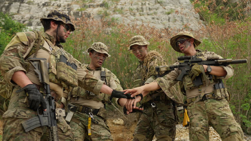 A diverse team of soldiers in full combat gear stacks hands together, demonstrating unity and strategizing for a military operation in a rugged environment.