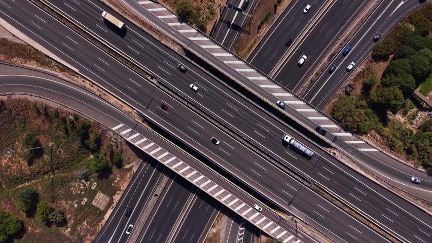 Top-down view of a multi-lane highway interchange with cars, trucks and a bus moving across elevated overpasses surrounded by sparse greenery