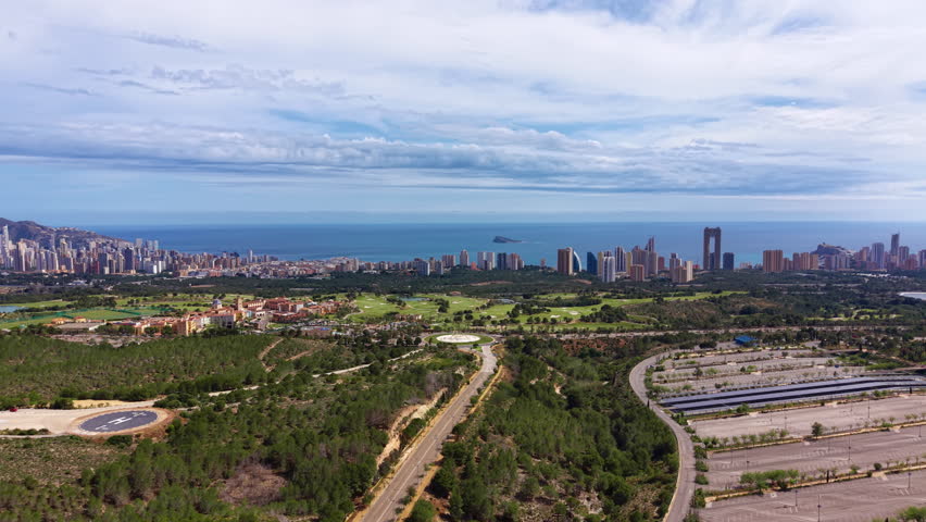 Panoramic view of Benidorm, Spain, showing skyscrapers along the Mediterranean coastline, a golf course, winding roads, solar panels, and lush greenery under a cloudy sky