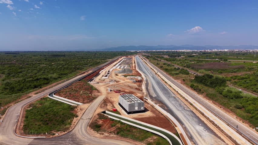 Highway construction site with partially completed road lanes, exposed earthworks, and concrete foundations stretching toward distant mountains under a clear blue sky