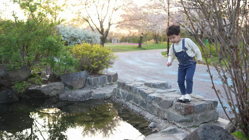 Child leaps across stone bridge with joy. Sunlight glows soft over garden pond. Pink blossoms frame serene outdoor scene. Perfect for nature, family, or childhood adventure shots