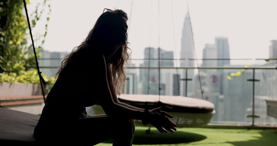 A pensive woman in silhouette sits on a rooftop swing, contemplating the city skyline and urban life.