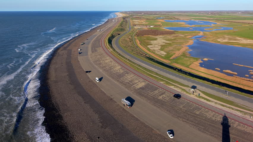 Aerial drone view of a coastal landscape featuring a long curved dike separating the open sea from inland terrain. A paved road runs along the top of the dike, with several vehicles traveling or parked on the route. Ocean waves break along the rocky shoreline on the seaward side. Inland, multiple parallel roads and cycling paths extend across green fields interspersed with ponds, wetlands, and narrow strips of vegetation. The environment includes managed coastal infrastructure, transportation routes, and natural wetland areas visible across the landscape. The scene highlights the integration of coastal engineering, land management, and human activity within a protected shoreline region under clear daylight.