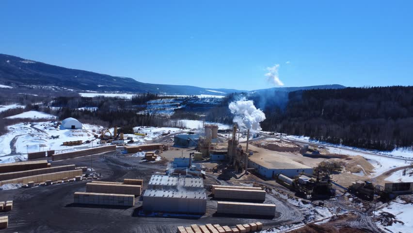 Active sawmill viewed from above, featuring wood stockpiles, drying facilities, and industrial infrastructure in a cold Canadian setting.