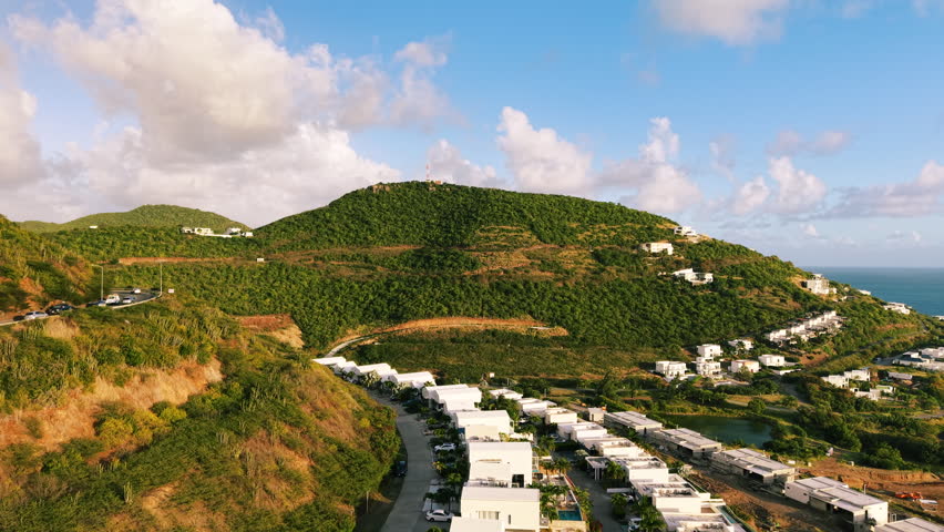 Aerial view of Saint Martin island in the Caribbean Sea. Green tropical hills with luxury villas and residential houses under a blue sky with clouds. Coastal neighborhood with white buildings along the road near the ocean coastline. Picturesque summer landscape.