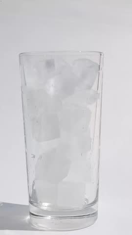 Water stream pours steadily into tall clear glass densely packed with ice cubes filling from bottom as liquid level rises and bubbles form around ice on white background.