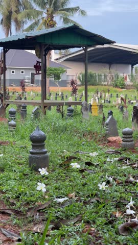 A peaceful Muslim cemetery with traditional tombstones or nisan on a green grassy ground with scattered white flowers.