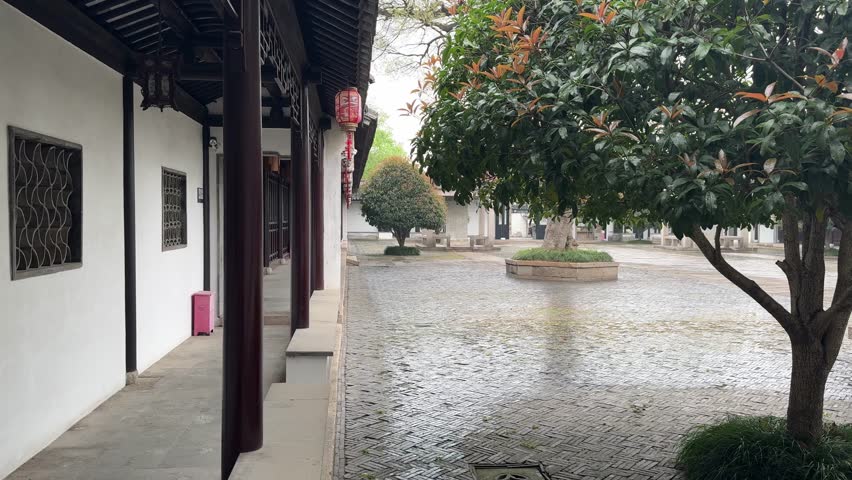 Serene traditional Chinese courtyard corridor with dark wooden pillars, white plaster walls, decorative lattice windows, and hanging red lanterns. Cobblestone plaza features raised circular tree plant