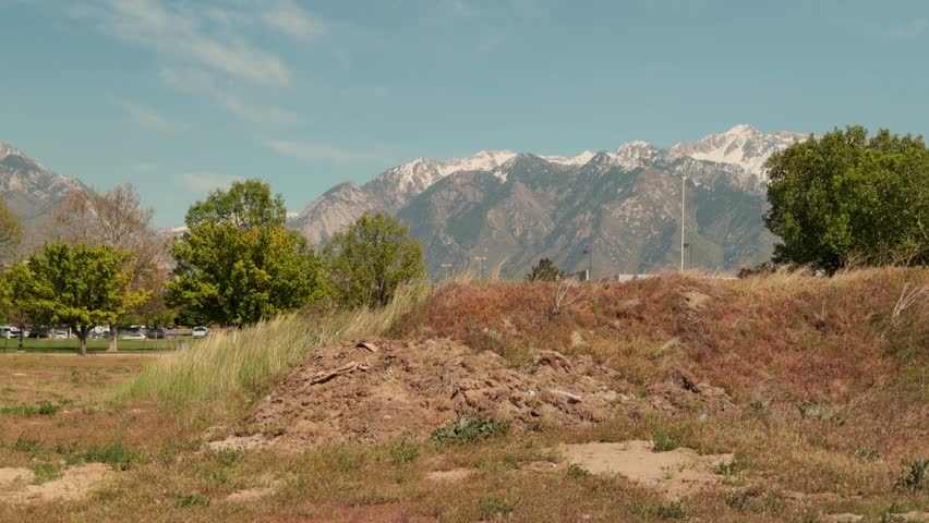 Static shot of dry hillside and scattered trees with distant snow covered mountain range under clear sky.