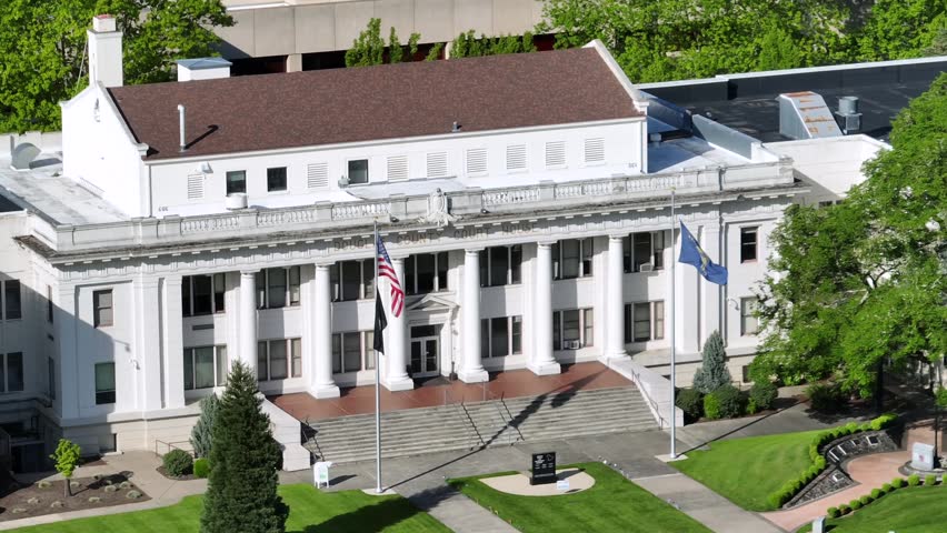 Historic white columned courthouse with American and Oregon state flags flanking grand entrance steps, manicured lawn and green trees in downtown Roseburg, Oregon, USA. Aerial drone view.