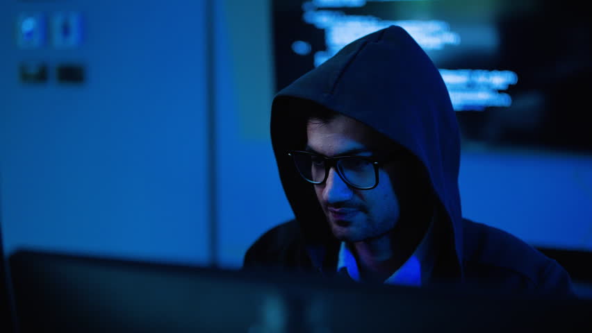 Male hacker in a black hoodie working on a computer in a dark room with blue neon light. He celebrates a successful cyber attack or coding breakthrough. Technology and cyber security concept.