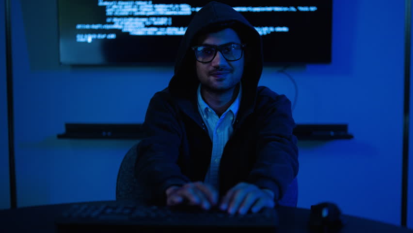 Male hacker in a black hoodie working on a computer in a dark room with blue neon light. He celebrates a successful cyber attack or coding breakthrough. Technology and cyber security concept.