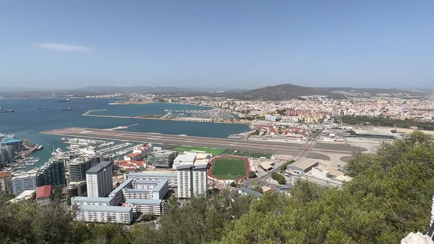 Wide angle shot from Rock of Gibraltar shows city skyline, marina, airport runway, blue bay, distant hills, bright daylight, clear sky, and lush foreground vegetation in sharp focus.