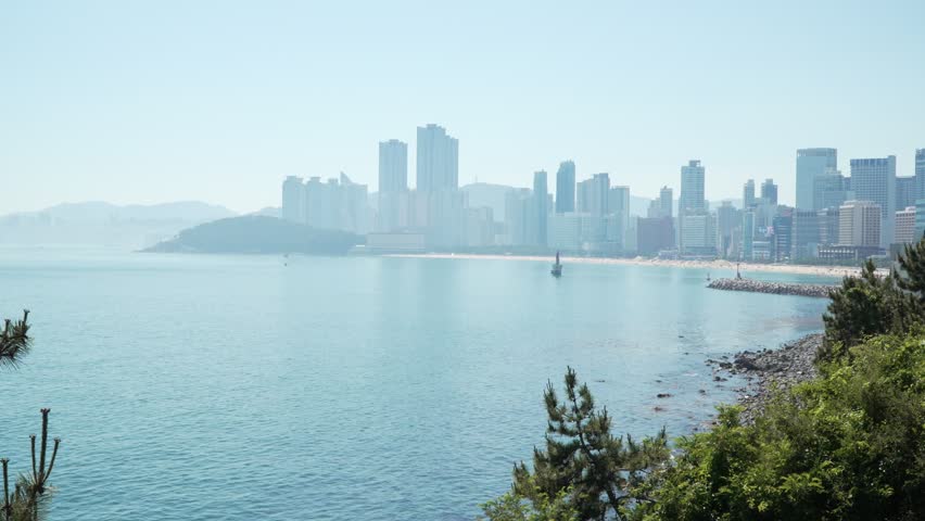 Hazy view across calm bay toward Haeundae beach and skyline of high-rise towers seen from Haeundae Blue Line Park in Busan South Korea