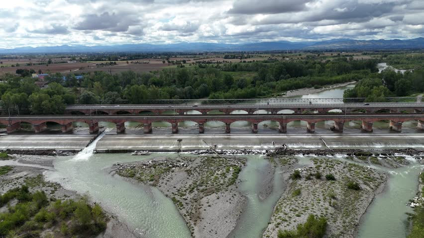 Parallel road and railway bridges cross the Trebbia River near Rottofreno, Piacenza, Emilia-Romagna, Italy, braided channels and gravel bars, with vehicle traffic visible on the roadway, slow push in