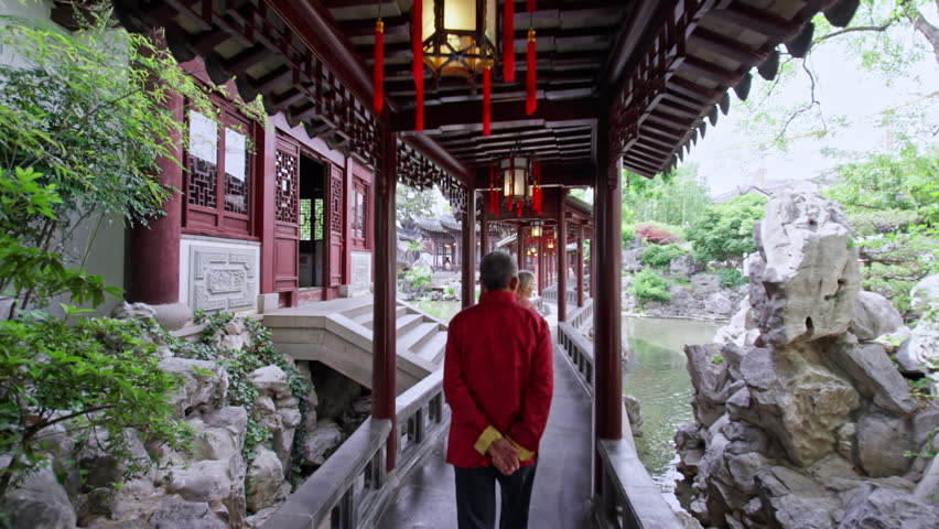 An elderly man in a red jacket strolls through a covered walkway in a traditional Chinese garden. Ideal for cultural documentaries or travel features.