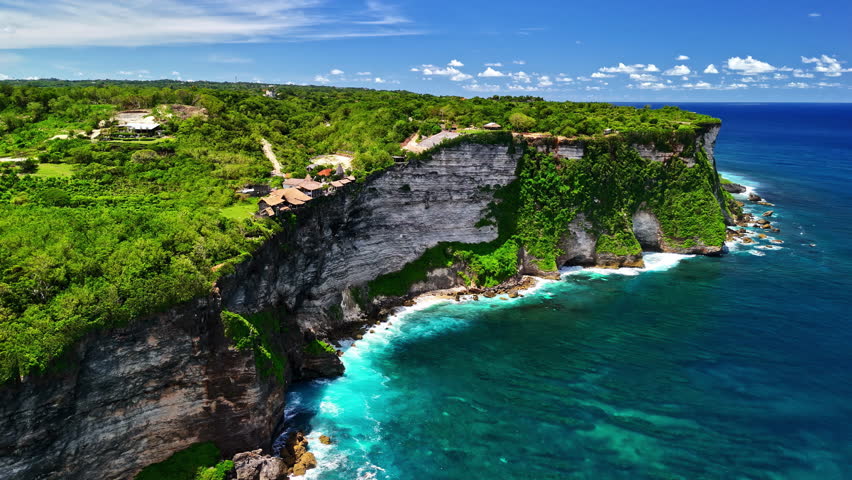 Aerial view of towering limestone sea cliff covered in green jungle with turquoise ocean and crashing waves near Nyang Nyang Beach in Uluwatu Bali Indonesia