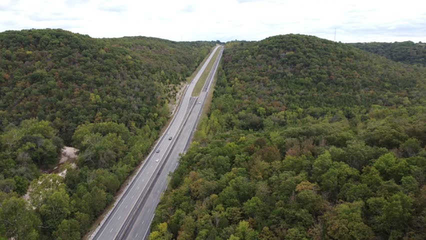 Cinematic aerial footage showcasing a highway stretching through the rolling hills and dense forests of Busiek State Forest and Wildlife Area in southwest Missouri.