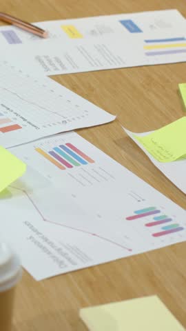 Vertical video: Panning camera showing charts on table in office with pen, sticky notes for review. Round, wooden, conference, reports, data, analysis, vertical video