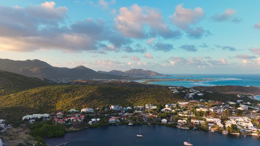 Drone aerial view of Oyster Pond bay on the border of Sint Maarten and Saint Martin. Tropical island landscape features luxury coastal houses and boats anchored in the calm harbor. Green hills and blue ocean waters stretch toward the horizon under a cloudy sky. Scenic destination on the Caribbean coast perfect for travel, real estate, and vacation themes.