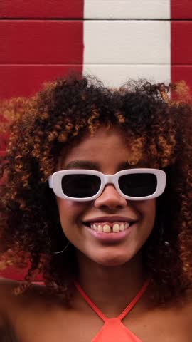 Joyful young woman with curly afro hair wearing stylish sunglasses and smiling happily at the camera against a striped red and white wall, expressing positivity and confidence in a city environment