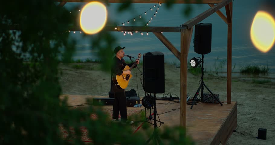 Male acoustic guitarist performing on a small beachfront stage at dusk