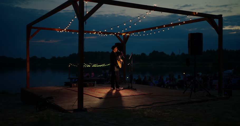 Solo musician performing acoustic set on lakeside wooden stage at dusk