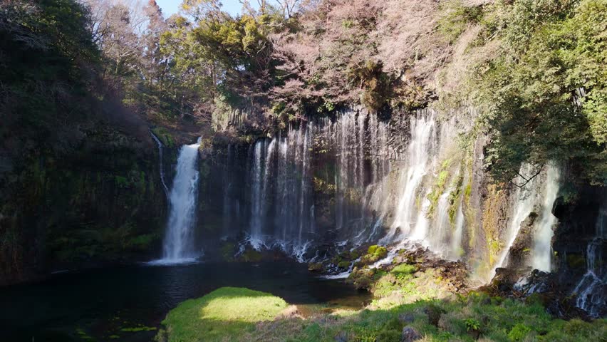 Drone video ascending near a spectacular wide curtain waterfall with silky water flowing down moss-covered rocks.