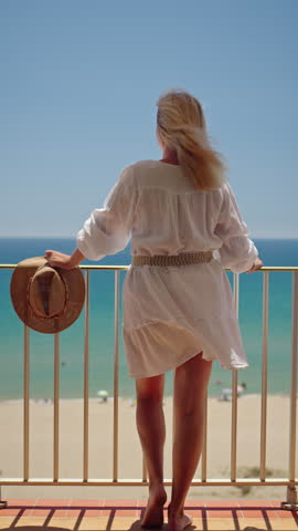 Woman in white summer dress standing on balcony with straw hat and looking at turquoise sea during sunny beach vacation.