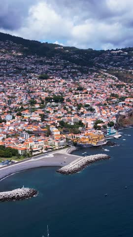 Funchal coastline in Madeira, Portugal, featuring curved breakwaters, hillside housing, marina scenery, and Atlantic Ocean views, vertical aerial cinematic pan