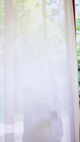 Soft light filters through sheer curtains behind a woman. She wears a modest hijab and dark clothing. Her gentle gaze suggests calm serenity. The scene feels peaceful and intimate