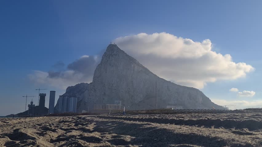 Low angle, wide shot of Gibraltar’s iconic rock formation from a sandy beach. Dramatic white cloud hangs over the peak, with bright daylight and crisp sky enhancing the striking scene.