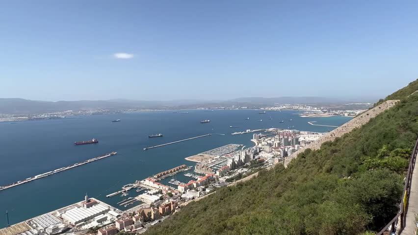 Drone video captures a sweeping panoramic view from the Rock of Gibraltar, showing the coastal city, vibrant harbor, cargo ships on vivid blue water, and sunlit landscape under a clear sky.