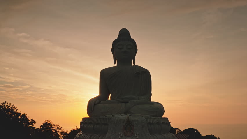 Big Buddha silhouette - Phuket famous landmark sitting on hillside, serene landscape during vibrant orange sunrise, peace and spiritual contemplation in Thailand. Aerial drone flight zoom out