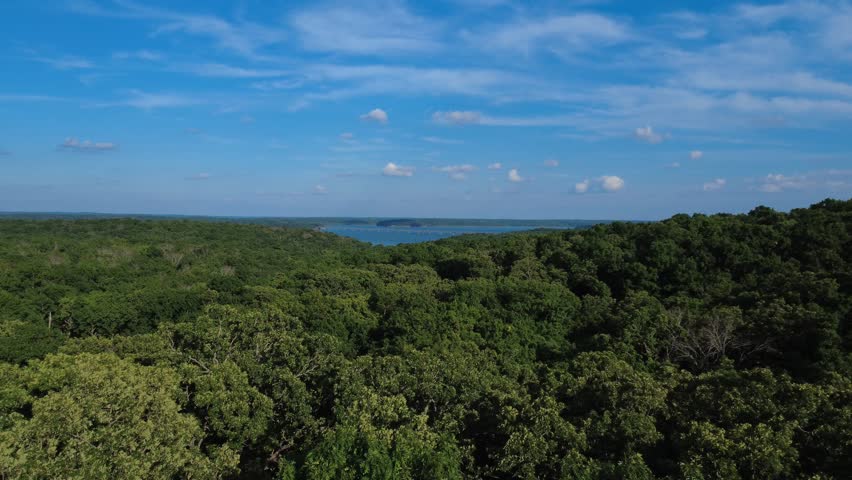 Drone footage over Ozark forest near Stockton Lake, Missouri, with distant lake views, a long bridge, blue sky, and wooded ridgelines.