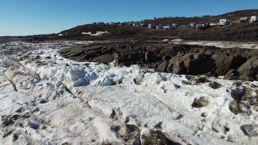 Drone shot tracking the stark line where a thick white ice foot meets the dark, snow-patched rocky shoreline and houses of Iqaluit.