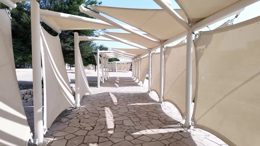 A perspective view of a paved outdoor walkway covered by a series of white fabric tension structures providing shade in a sunny park setting.