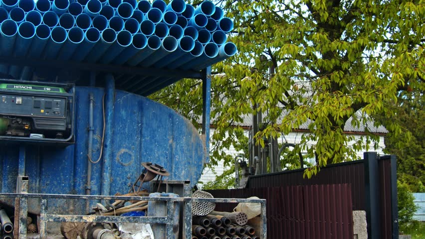 Workers prepare drilling equipment for a water well. The site shows various tools and blue pipes ready for use, with a tree and building in the background.