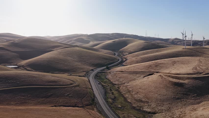 A drone view captures the Altamont Pass landscape, where wind turbines line the ridges and a curving road weaves through the hills, reflecting the region’s blend of infrastructure and nature.