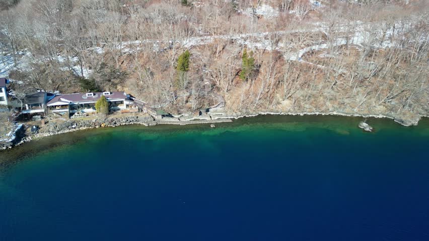 High-angle aerial shot tracking above a vibrant turquoise and blue lake shoreline with a cozy lakeside house hidden in winter woods.